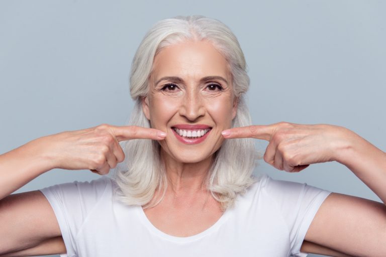 Older adult with white hair smiling broadly and pointing to their teeth with both index fingers, wearing a white shirt against a light blue background. Expression conveys confidence in dental health.