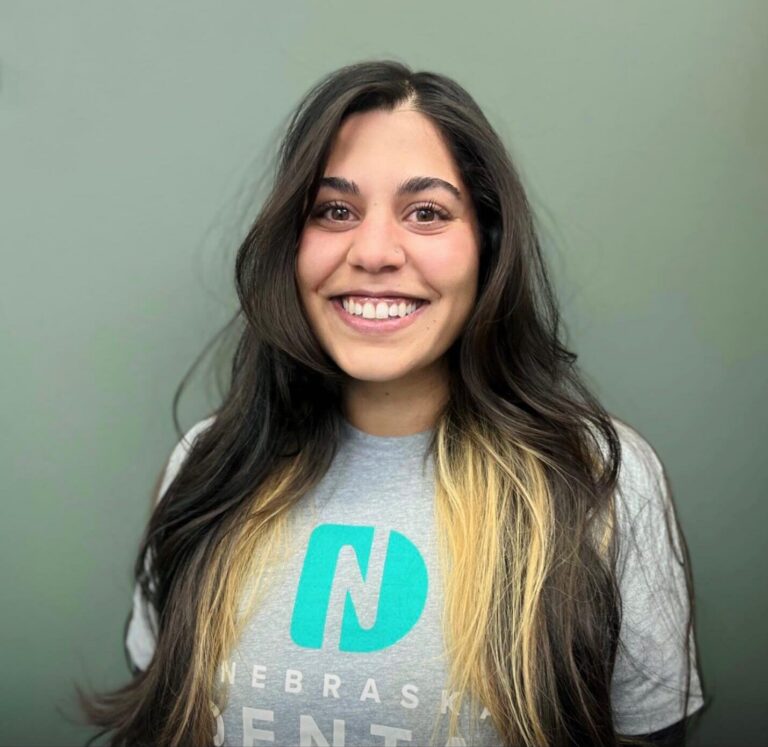 Headshot of Dana a front office administrator wearing a shirt with a logo of Nebraska Dental, in a plain gray background.