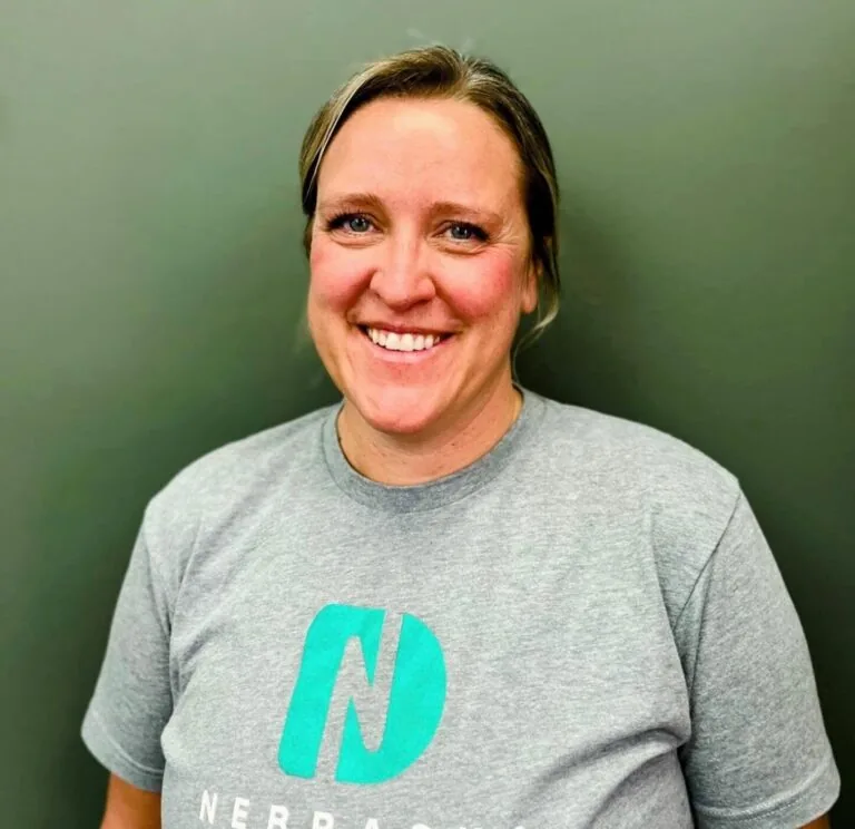 Headshot of Janel a dental hygienist wearing a shirt with a logo of Nebraska Dental, in a plain gray background.