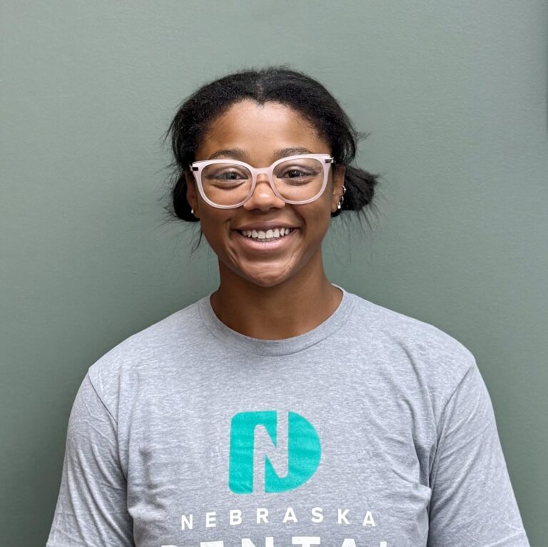 Jeana a dental assistant with two buns wearing light gray “Nebraska” shirt and pink glasses, standing against plain green wall.