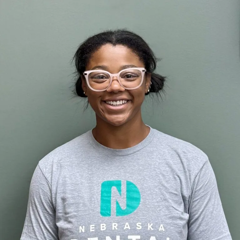Jeana a dental assistant with two buns wearing light gray “Nebraska” shirt and pink glasses, standing against plain green wall.