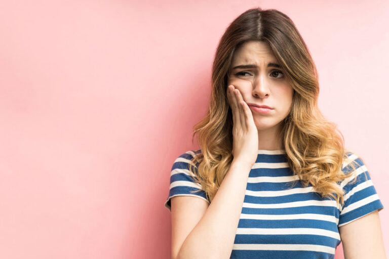A young woman with long wavy hair wearing a blue-and-white striped shirt stands against a pink background, pressing her hand to her cheek with a pained expression, suggesting dental discomfort.