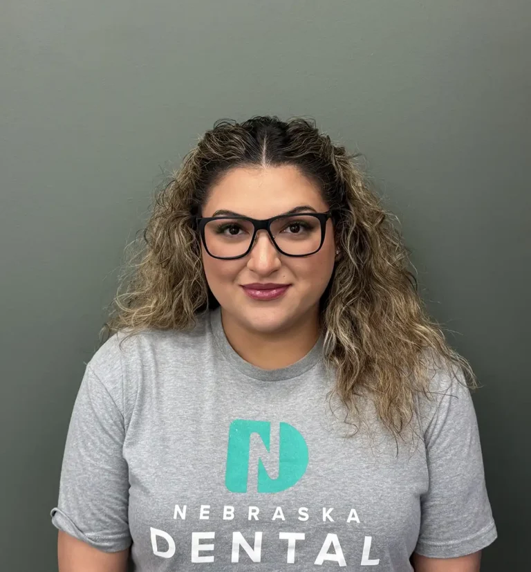 Jesyy wears glasses and a Nebraska Dental Center t-shirt, smiling warmly against a simple background while representing Nebraska Dental Center.